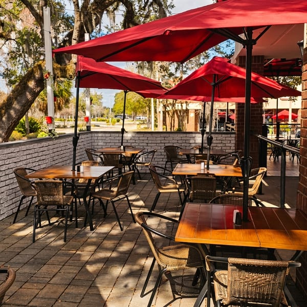 Outdoor restaurant patio with wooden tables, wicker chairs, red umbrellas, and brick walls under trees.