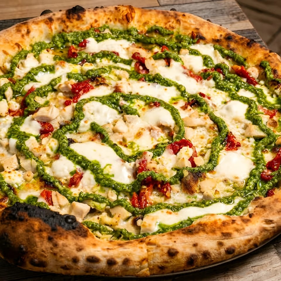 A whole pizza with spiral pesto, chicken, mozzarella, and sun-dried tomatoes on a wooden table.