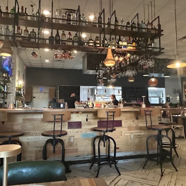 Restaurant interior with a rustic bar, bottles on shelves, hanging lights, and people dining.