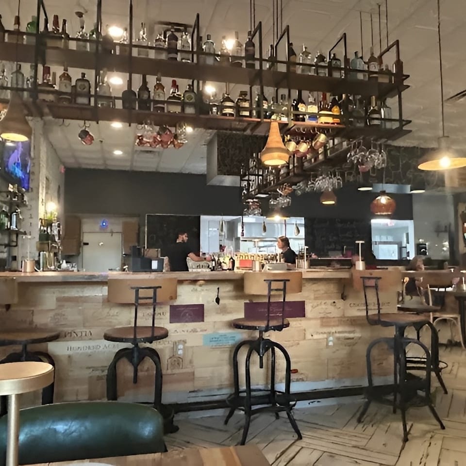 Restaurant interior with a rustic bar, bottles on shelves, hanging lights, and people dining.