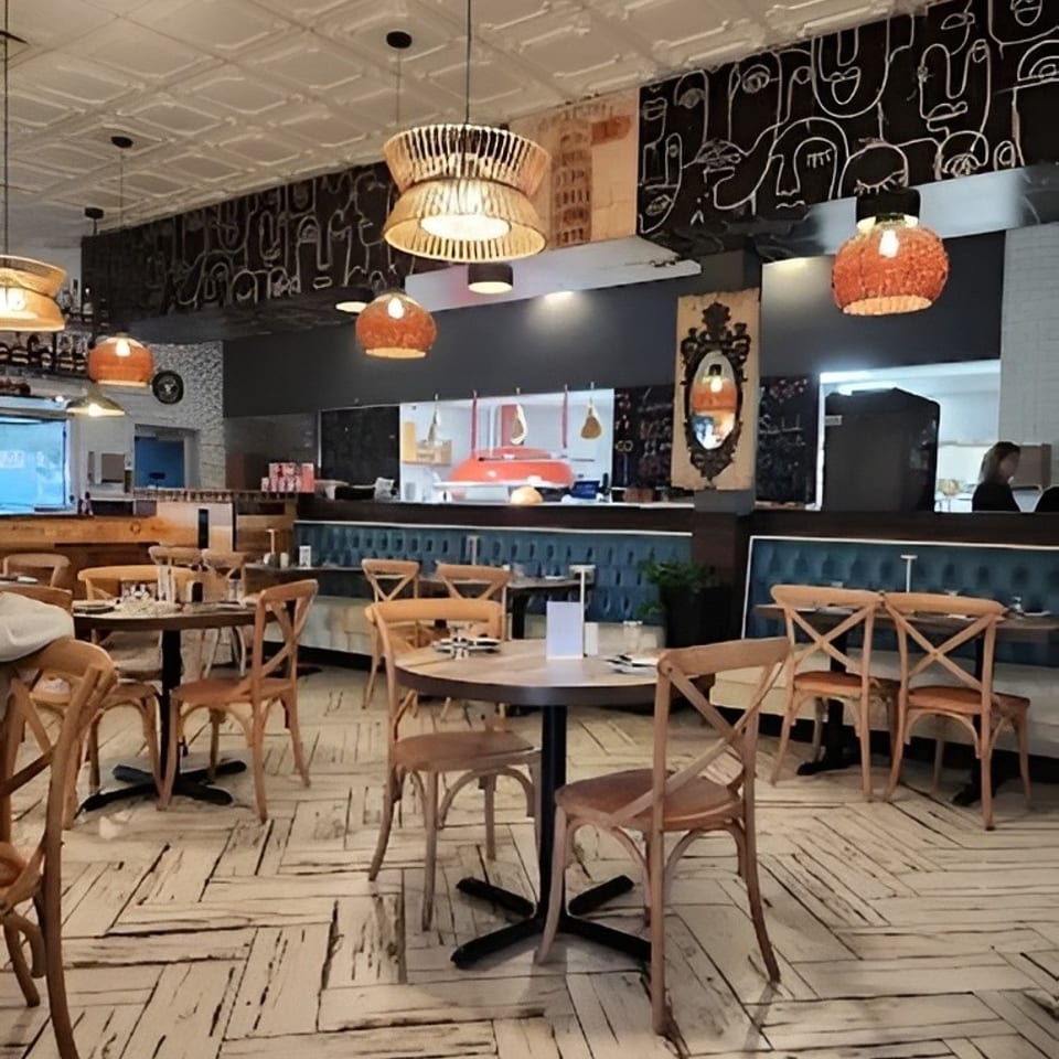 Cozy restaurant interior with wooden tables, chairs, unique lighting, and an open kitchen.